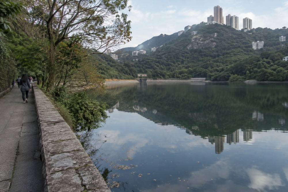 A stone path beside a tree-lined reservoir with hills rising behind it