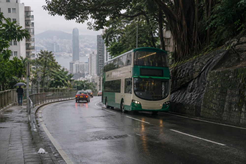 Double-decker bus on a winding road climbing through green hillsides above Hong Kong towers