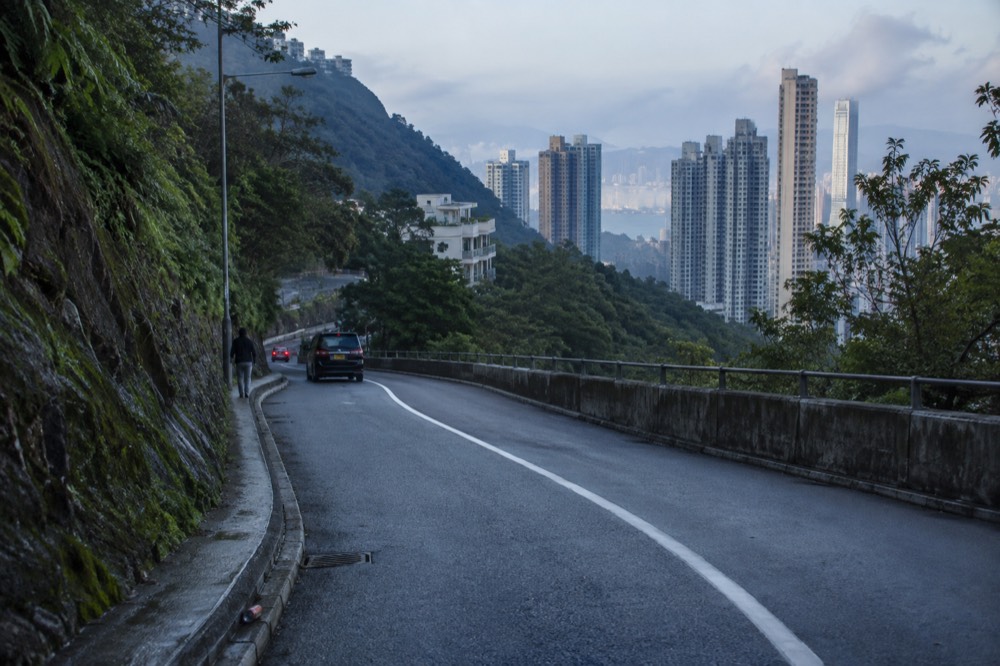 A descending road curving down a green hillside toward distant high-rise apartment blocks